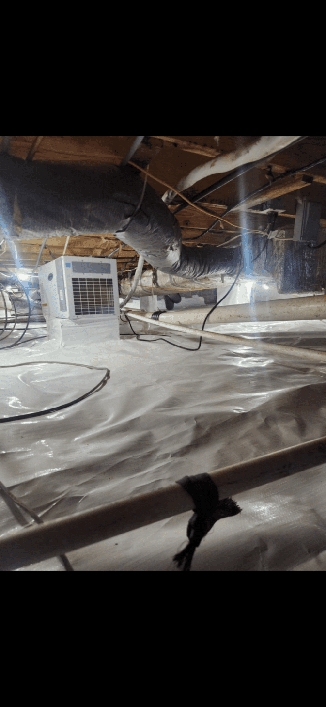 A crawl space with a white vapor barrier covering the ground, black ducts overhead, exposed wooden beams, and a white dehumidifier unit installed.