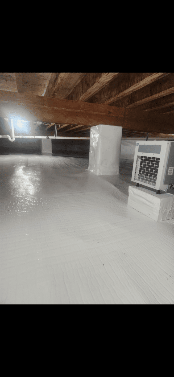 A crawl space with a white vapor barrier covering the floor and walls, exposed wooden beams above, and an HVAC unit installed on the right side.