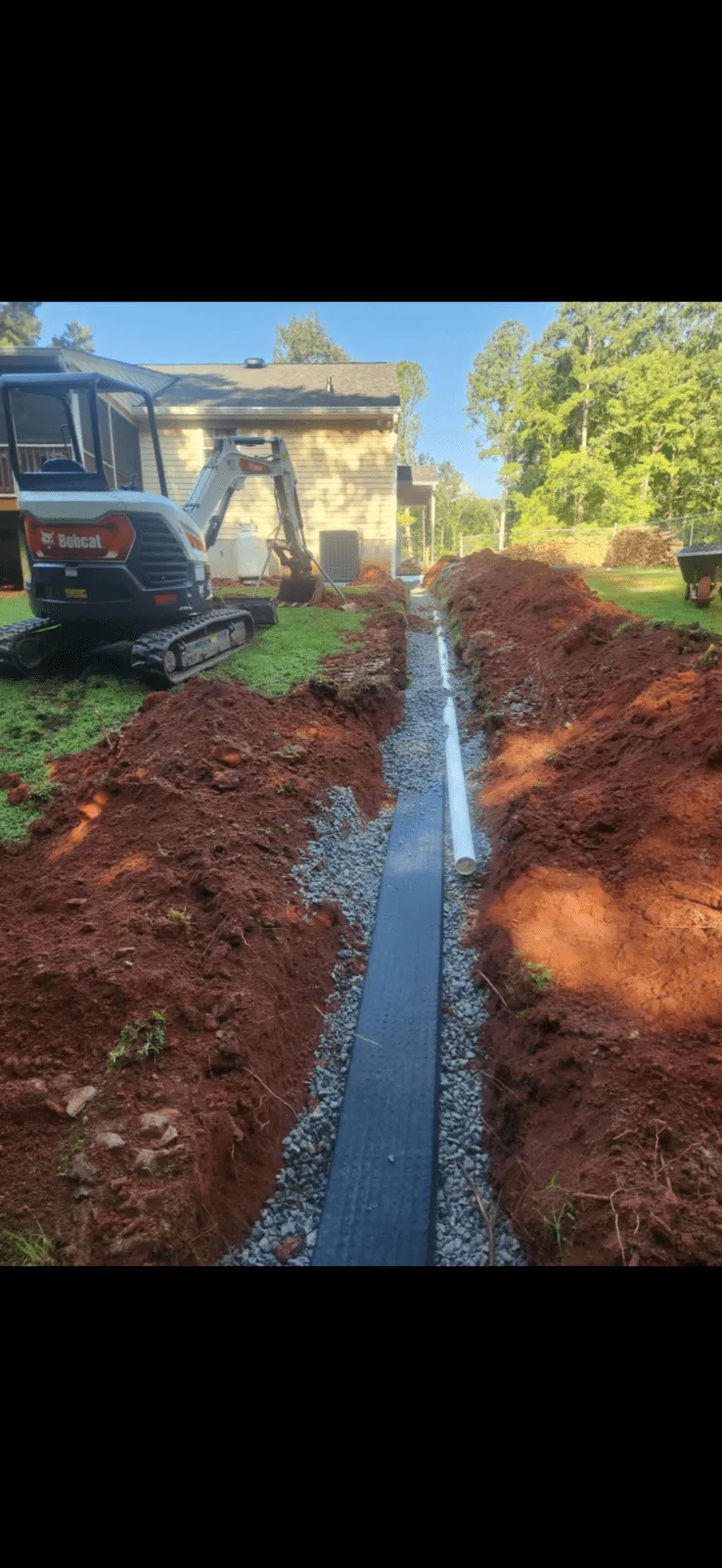 A trench with gravel, a black drainage mat, and a white PVC pipe runs through a yard; a mini excavator and wheelbarrow are nearby, with a house in the background.
