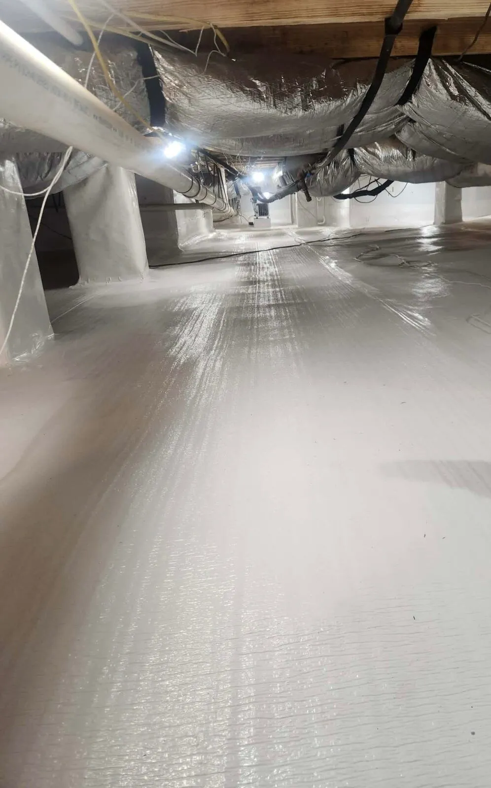 Clean, sealed crawl space with white vapor barrier lining the floor and walls, overhead pipes, wires, and insulation visible along the ceiling—showcasing professional waterproofing service for lasting protection.