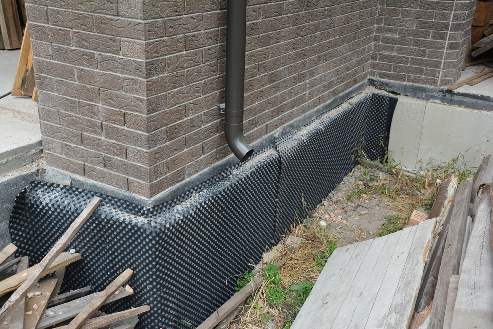 Close-up of a building foundation with a black drainage membrane installed, next to a downspout and surrounded by construction debris and wooden planks.