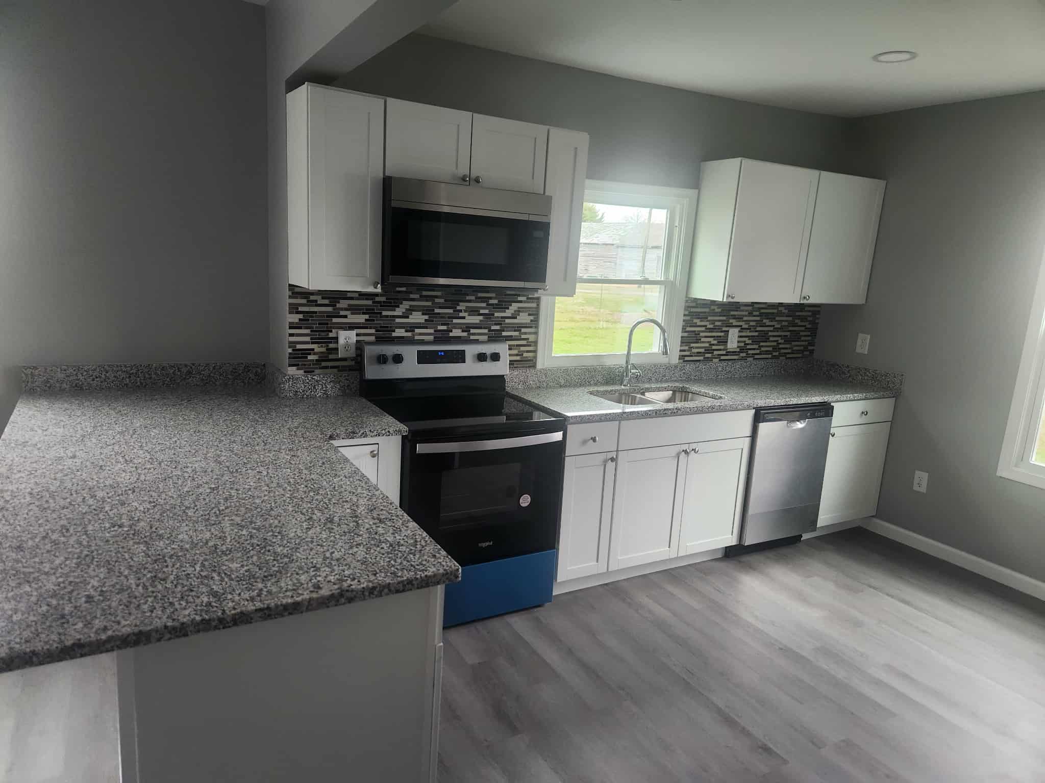 Modern kitchen with white cabinets, gray granite countertops, stainless steel appliances, mosaic tile backsplash, and light wood flooring. A large window provides natural light.