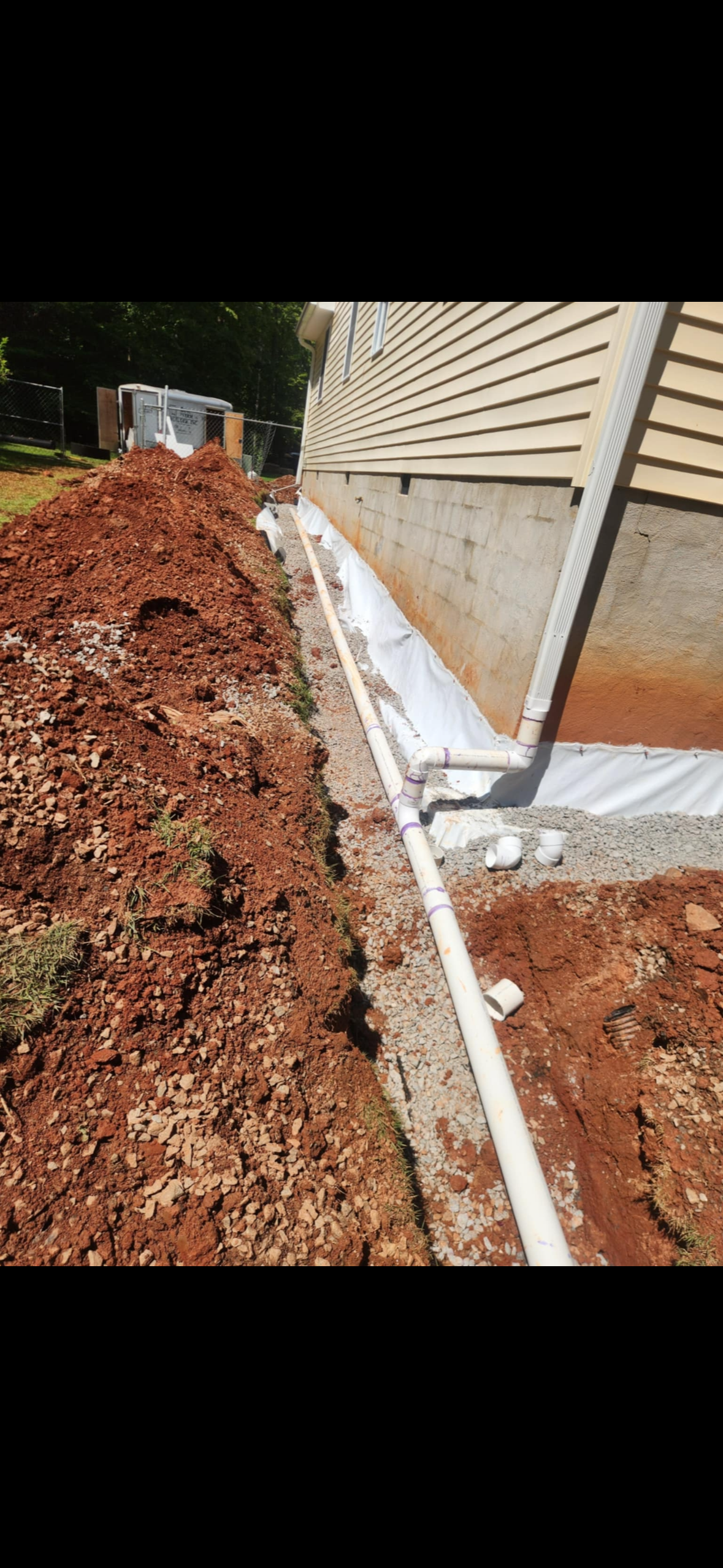 White drainage pipes are installed alongside a house foundation with exposed soil, gravel, and a trench running parallel to the building.