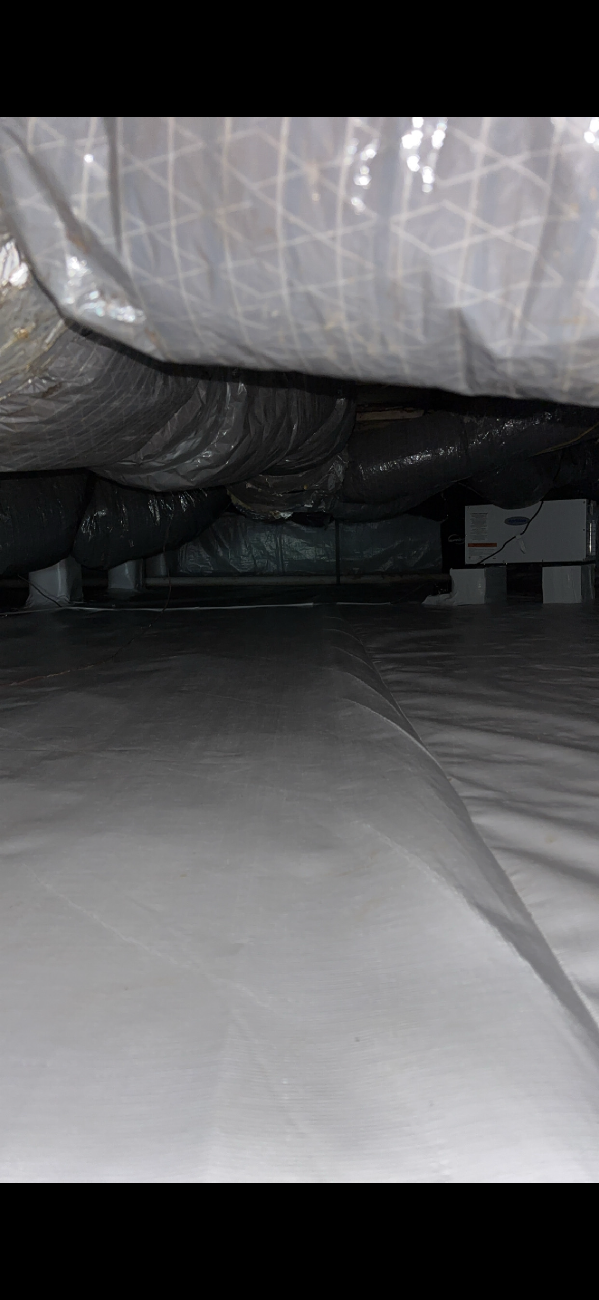 A photo of a crawlspace with plastic vapor barrier on the floor, insulated air ducts on the ceiling, and equipment along the sides.