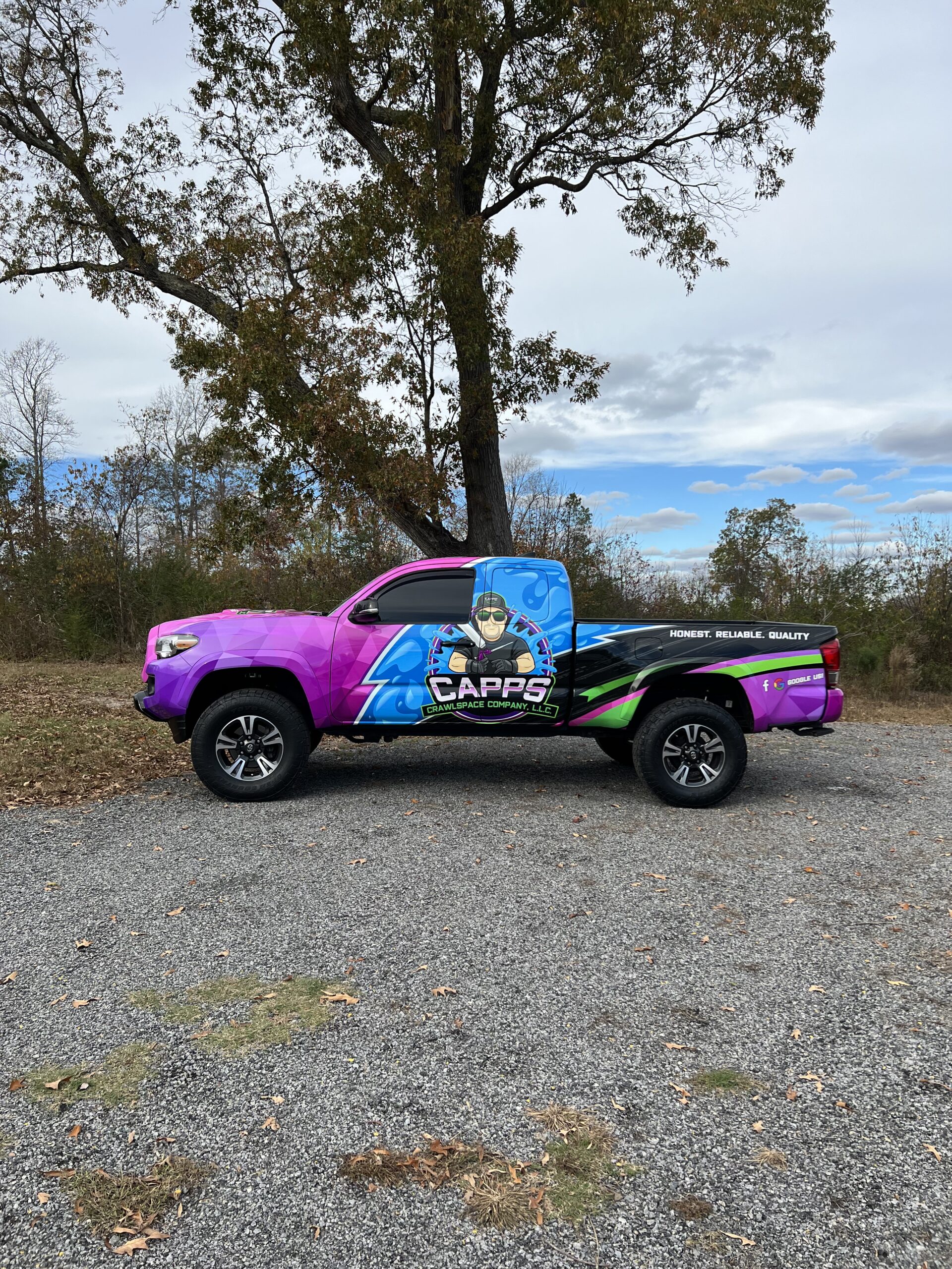 A pickup truck with a colorful, custom wrap featuring a cartoon character and the name Capps parked on gravel near trees and a cloudy sky.